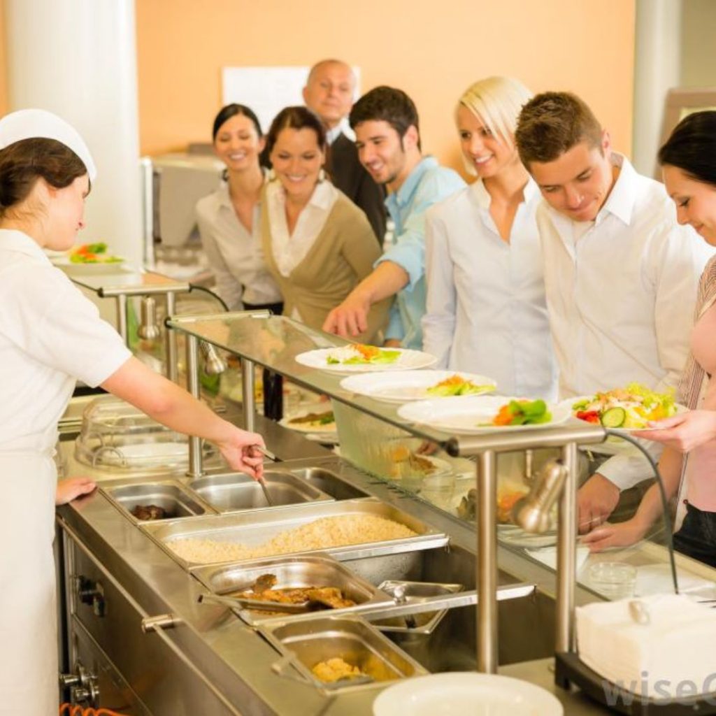 A group of people in business attire from the Department of Education stand in line at a cafeteria-style buffet. A server in a white uniform assists them, and the counter is lined with trays of food, including rice and vegetables, creating a clean and bright atmosphere.