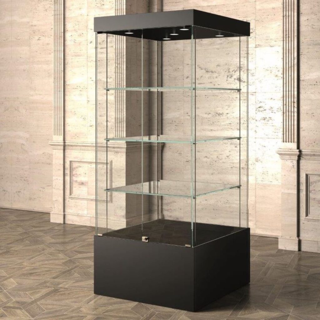 A tall, transparent glass display case with five shelves and a black base stands on a wooden floor in front of a textured beige wall. Perfect for showcasing educational awards, the case is empty, with prominent spotlights at the top to highlight future achievements.