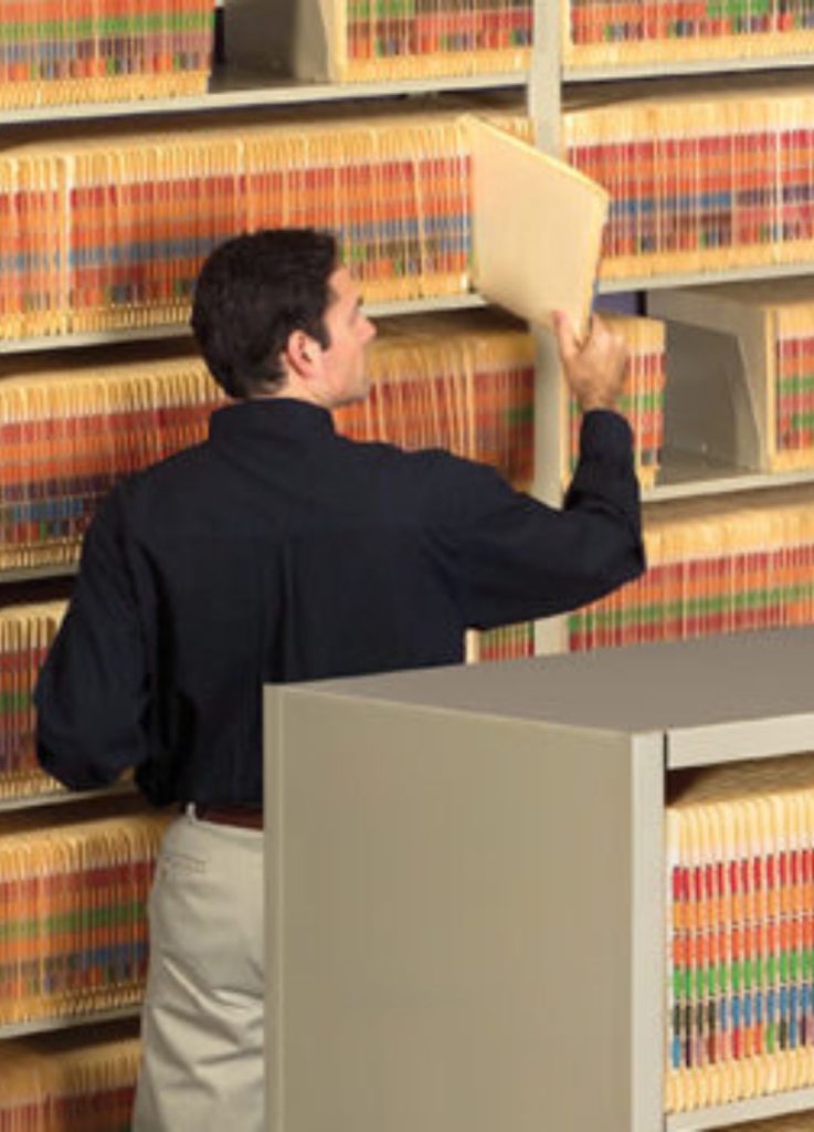 A person in a black shirt and beige pants places a file into a filing system on heavy-duty shelving units. The shelves, filled with color-coded folders, suggest an organized office or records environment.
