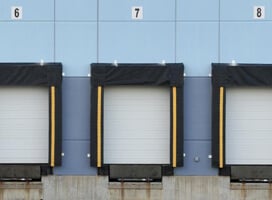 Three loading dock doors, numbered 6, 7, and 8, set against a blue industrial building wall. The docks have black padding, and the doors are closed—perfect for showcasing Newcastle Systems Brochures on portable power systems.