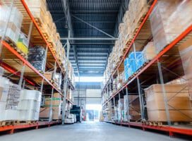 Wide-angle view of a warehouse interior with high shelves filled with stacked boxes and goods. The aisles are clear, and a forklift is visible in the background near an open loading bay. Mobile Powered Workstations from Newcastle Systems complement the bright lighting, enhancing productivity solutions throughout.