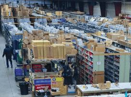 A large warehouse interior with stacks of boxes and shelves filled with various items. Two people stroll down an aisle among organized products. A workspace, featuring Newcastle Systems Brochures and portable power systems for enhanced productivity solutions, is visible in the foreground.
