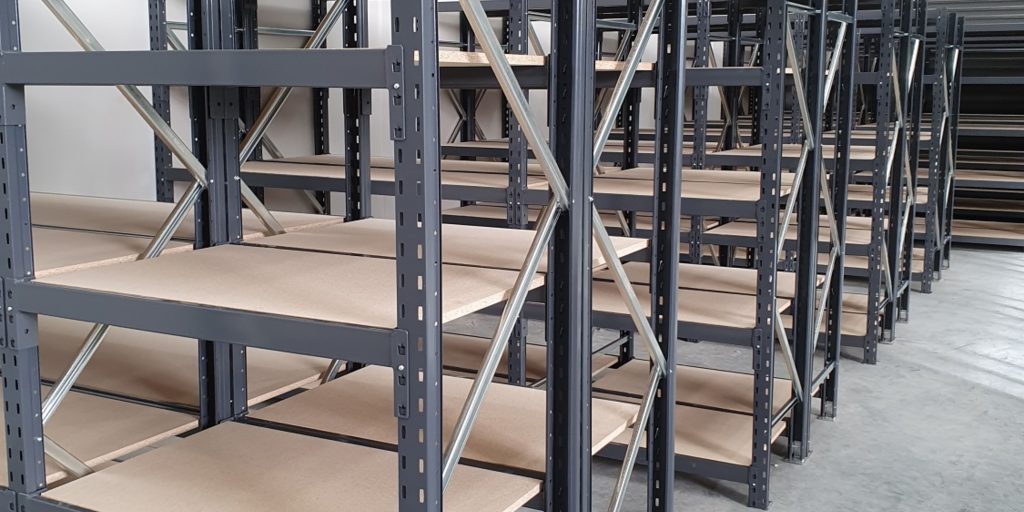 Rows of empty metal shelving units in a Jaken Images warehouse. The shelves have a wooden base and are organized in a spacious, well-lit area with a concrete floor.