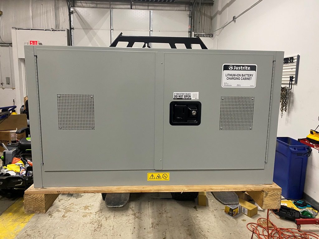 A large gray cabinet, labeled Lithium-Ion Battery Charging Safety Cabinet, stands on a wooden platform in an industrial setting. Equipped with the ChargeGuard™ Containment System, it features ventilation panels and a central black handle. Various tools and equipment are scattered around.