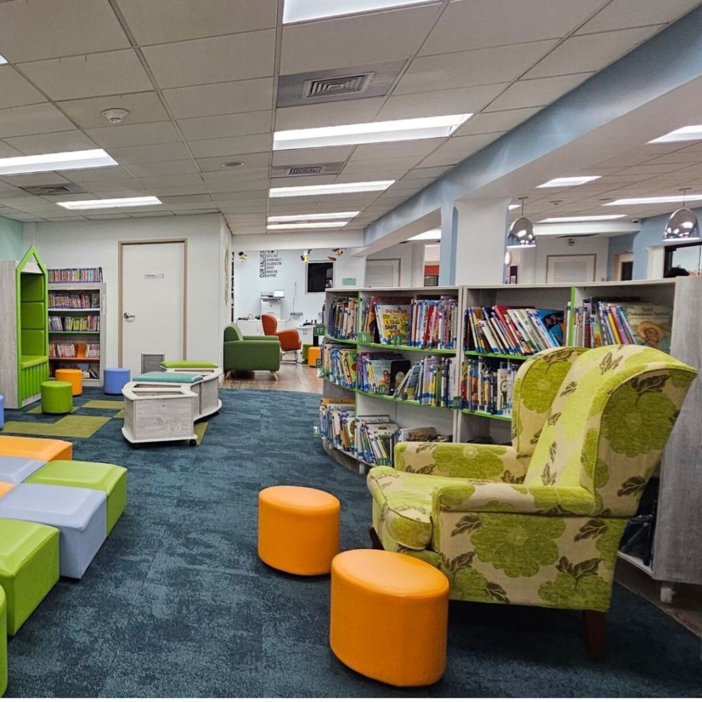 A cozy library interior in the department of education features a large floral armchair, orange stools, and colorful seating cubes on a carpeted floor. Bookshelves filled with childrens books line the walls, with low tables and green chairs scattered around.