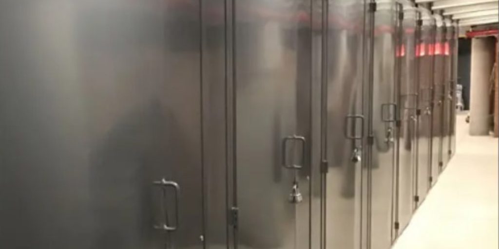 A row of large, dark-colored server cabinets, meeting top locker specifications with handles and locks, is lined up in a dimly lit room. Overhead piping and a concrete floor are visible, suggesting an industrial or data center environment akin to those on the Best Lockers specifications page.