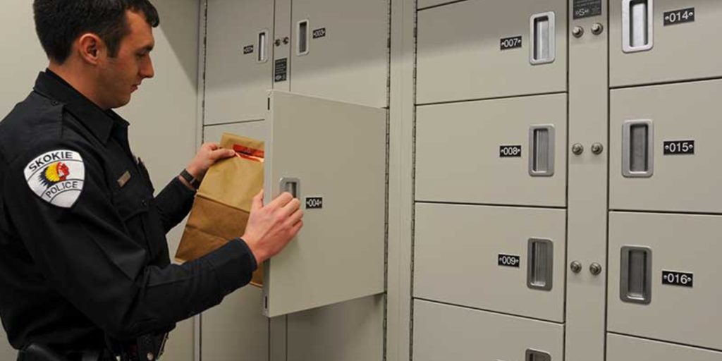A police officer from the Eagle Group places a brown paper bag into a numbered storage locker at the facility. The lockers are light gray, each labeled with unique numbers. The officers black uniform features a prominent patch on the sleeve.