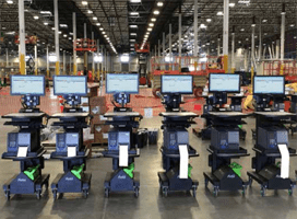 Several industrial mobile workstations with monitors and keyboards are lined up in a spacious warehouse setting. The background reveals high ceilings, metal structures, and several stacked materials, showcasing Newcastle Systems Brochures’ innovative productivity solutions.