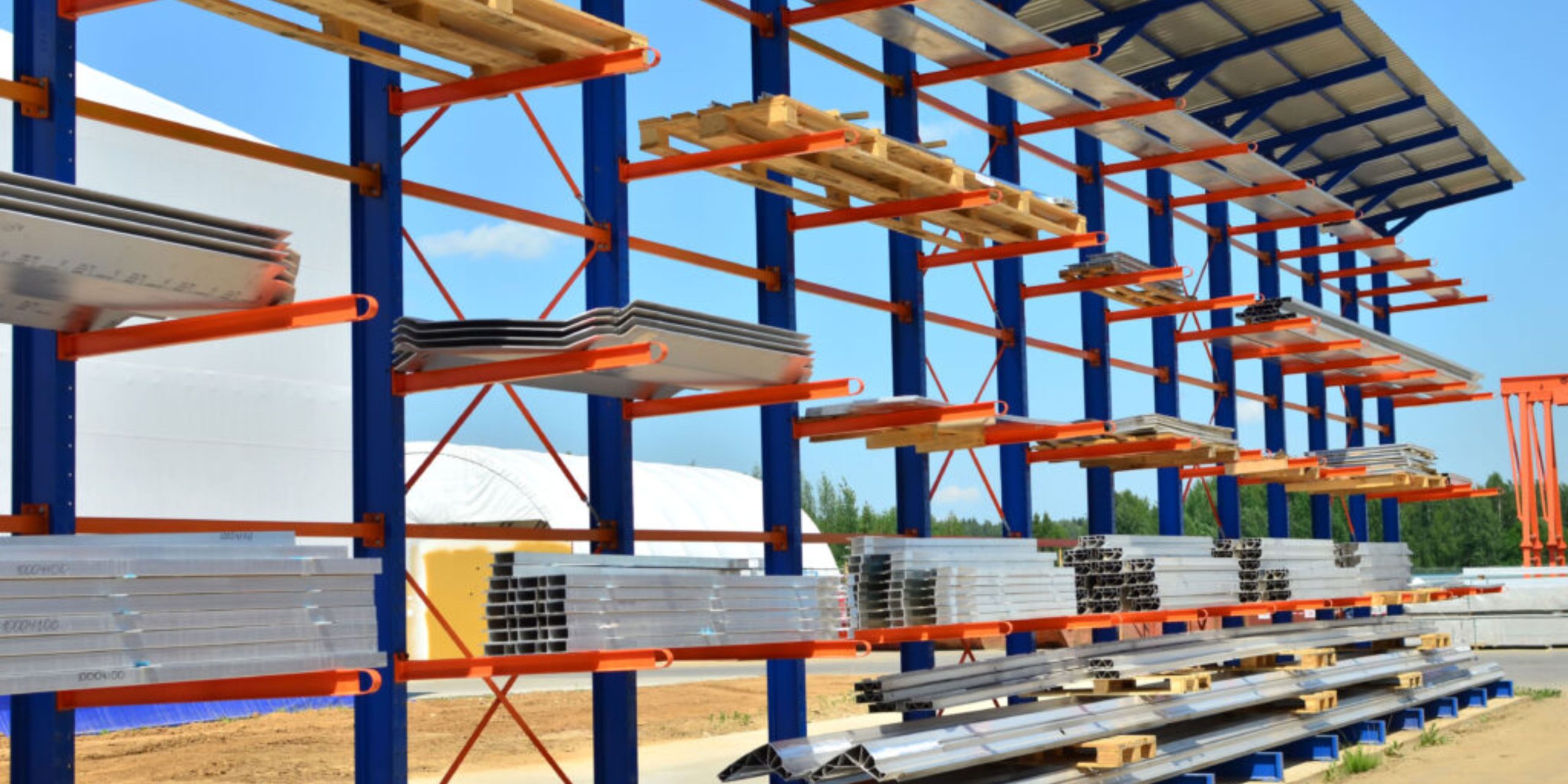 A shelving structure outdoors features stacked metal sheets and wooden planks under a metal roof, efficiently maximizing floor space. Supported by orange and blue beams, it stands against a backdrop of a white tent and clear blue sky.