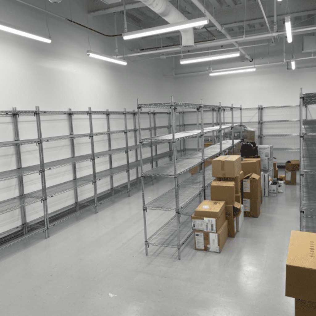 A room with metal shelving units lines the white walls, mostly empty like a material handling space. Cardboard boxes of various sizes are scattered on the shiny gray floor and some rest on shelves. The ceiling features visible pipes and fluorescent lights, complementing the utilitarian atmosphere.