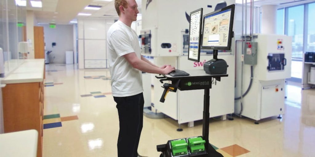 A person in a white t-shirt stands at an industrial mobile workstation with dual monitors in a bright office space. The workstation features a keyboard and green storage compartments, offering an ideal productivity solution. Office equipment is visible in the background.