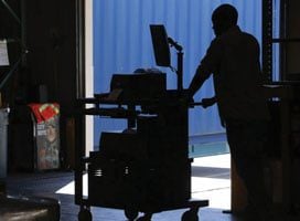 Silhouette of a person leaning on a cart with a monitor in a dimly lit space, showcasing Newcastle Systems portable power systems. Bright daylight and a blue industrial container are visible through an open door in the background, highlighting productivity solutions at work.