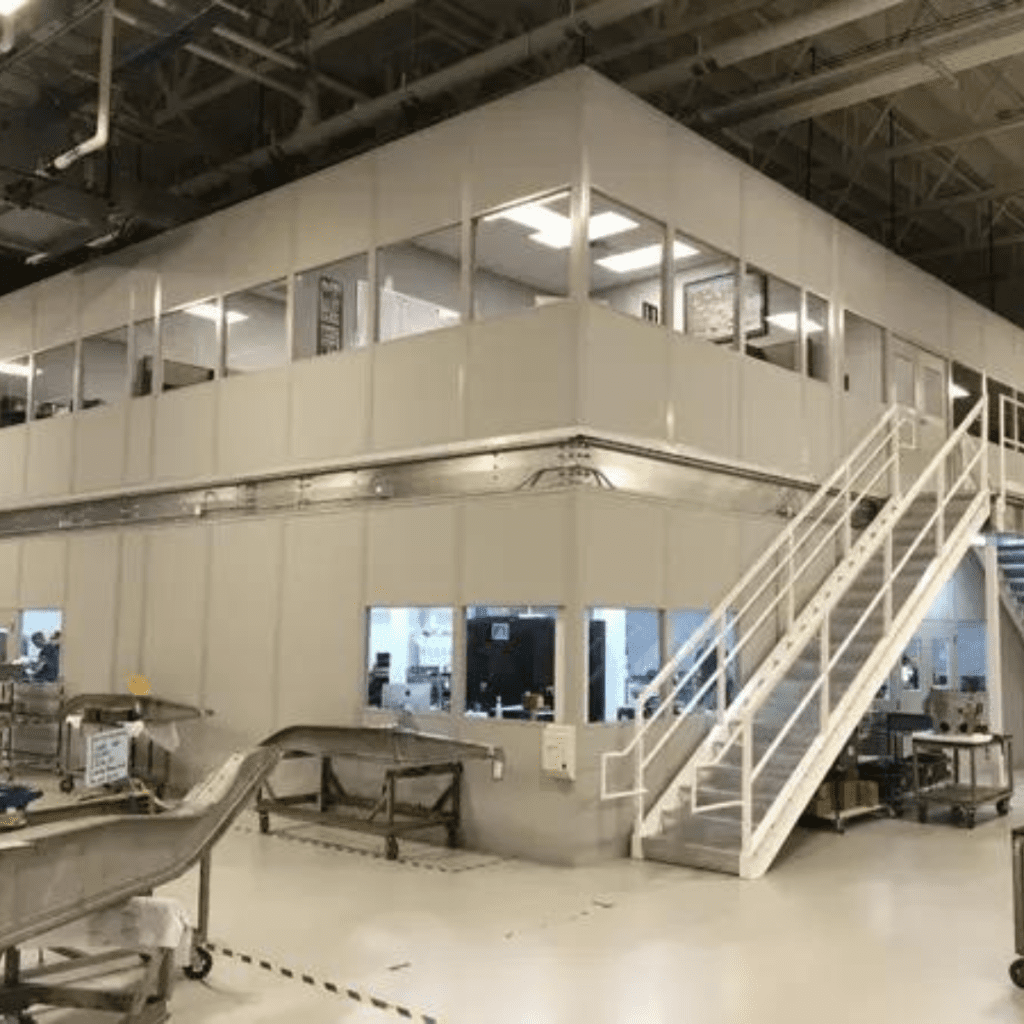 A large industrial room with a two-story white structure resembles a modern gallery. The upper floor, featuring windows and a staircase on the right, overlooks conveyor belts and material handling machinery below, all bathed in ample light.