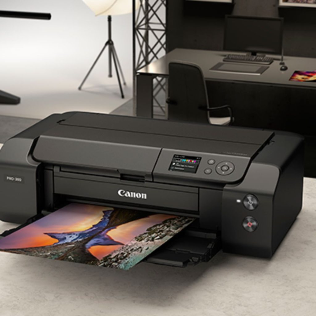 A Canon PRO-300 printer on a desk is printing a vibrant photo of a tree. The printers display shows colorful settings, perfect for educational projects. In the background, a workspace with a chair, desk, and photographic equipment sets the scene for creative learning.