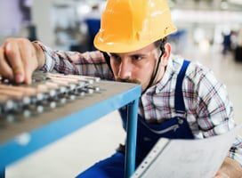 A man in a yellow hard hat and plaid shirt inspects machinery on the factory floor. Holding a document, he examines metal components on a blue workbench. The bustling industrial environment is enhanced by mobile powered workstations that boost productivity solutions for his team.