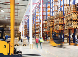 A bustling warehouse with tall shelves filled with stacked pallets buzzes with activity. Workers move efficiently next to a forklift transporting goods, aided by mobile powered workstations. The bright environment is illuminated by ample lighting, while a yellow structure stands prominently in the foreground.