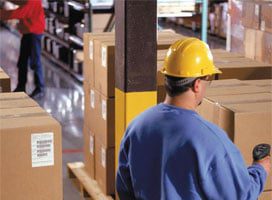 A worker in a blue shirt and yellow hard hat stands next to stacked cardboard boxes in a warehouse, holding a scanner on one of Newcastle Systems Industrial Mobile Workstations. More boxes fill the shelves in the background while someone in red is seen in the distance.
