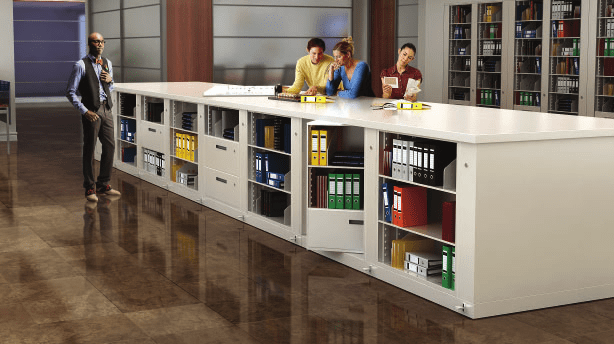 A group of people in a modern office space are engaged in storage planning, organizing files and folders on and inside a large white cabinet. The shiny brown tile floor complements the shelves filled with binders visible in the background.