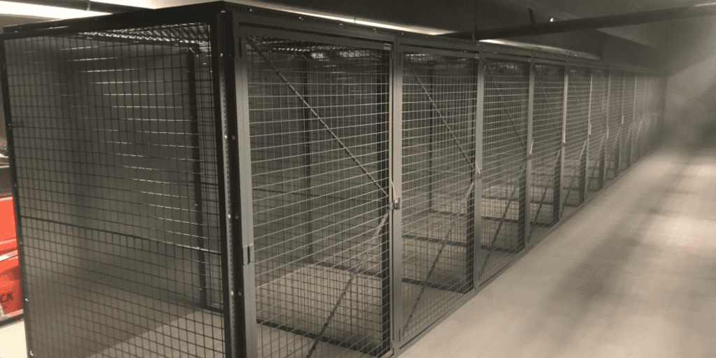 A row of spacious security cages with wire mesh walls stretches into the distance under artificial lighting, nestled in a dimly lit room. Aligned neatly, these cages are ideal for robust warehouse security and theft prevention solutions.