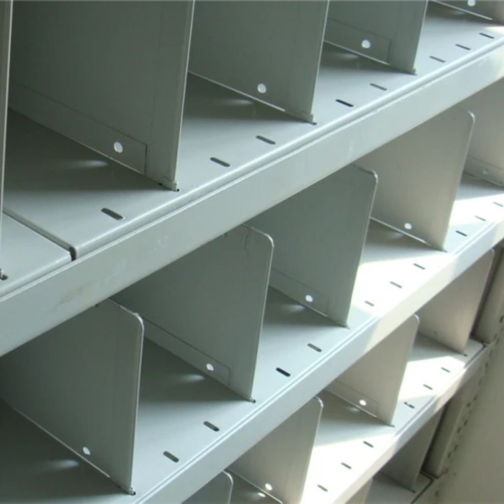 Close-up of empty metal shelves with vertical dividers, emblematic of industrial shelving solutions. The smooth, gray surface features small circular holes along the edges, suggesting modular or adjustable components. Lighting casts soft shadows on this heavy-duty shelving unit.