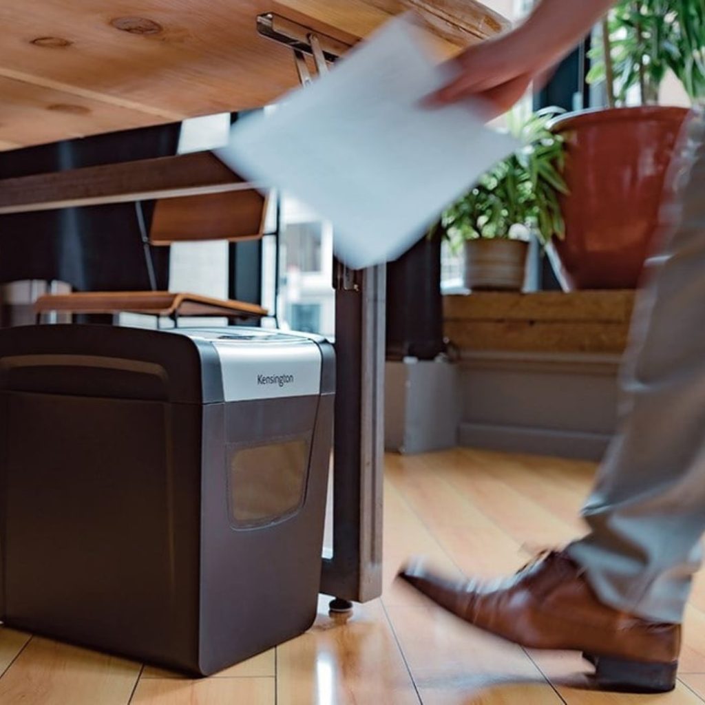 A person from the Department of Education walks toward a Kensington shredder under a wooden desk, clutching papers. The wooden floor and potted plants in the background complete this workspace setting.