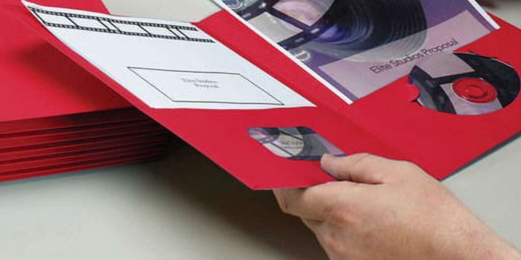 A person holds a red folder containing two CDs, a blank label, and a strip of film. The CDs are partially visible through cutouts in the folder. Smead Brochures stack neatly in the background, showcasing durable filing solutions that enhance office organization.