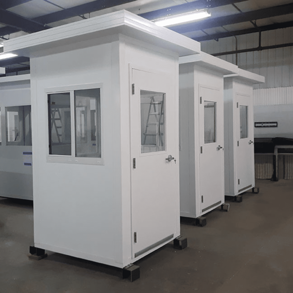 Three white security guard booths with windows and doors are aligned in a spacious, industrial warehouse setting. The booths, elevated slightly off the ground, overlook a material handling area, while a metal structure stands prominently in the background like an artistic gallery piece.