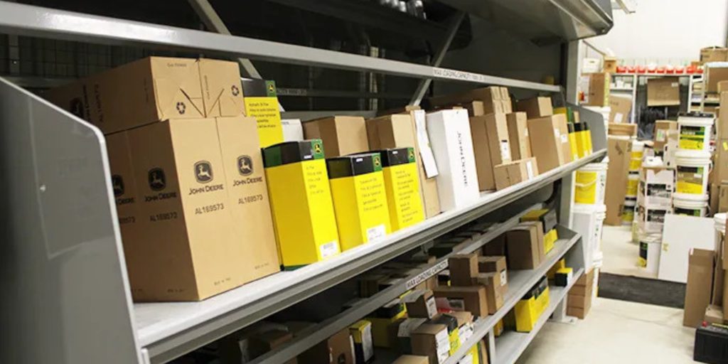 A storage room with sleek steel shelving holds neatly arranged cardboard boxes of various sizes, some featuring logos. In the background, more shelves and items are visible. The white floor enhances the organized look. Tri Boro Shelving Brochures are casually placed on a nearby utility cart.