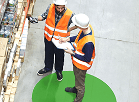 Two people wearing white hard hats and orange safety vests stand on a green circle in a warehouse aisle, accompanied by an Industrial Mobile Workstation. One holds a clipboard, the other a scanner, as they consult Newcastle Systems Brochures amidst shelves filled with boxes.
