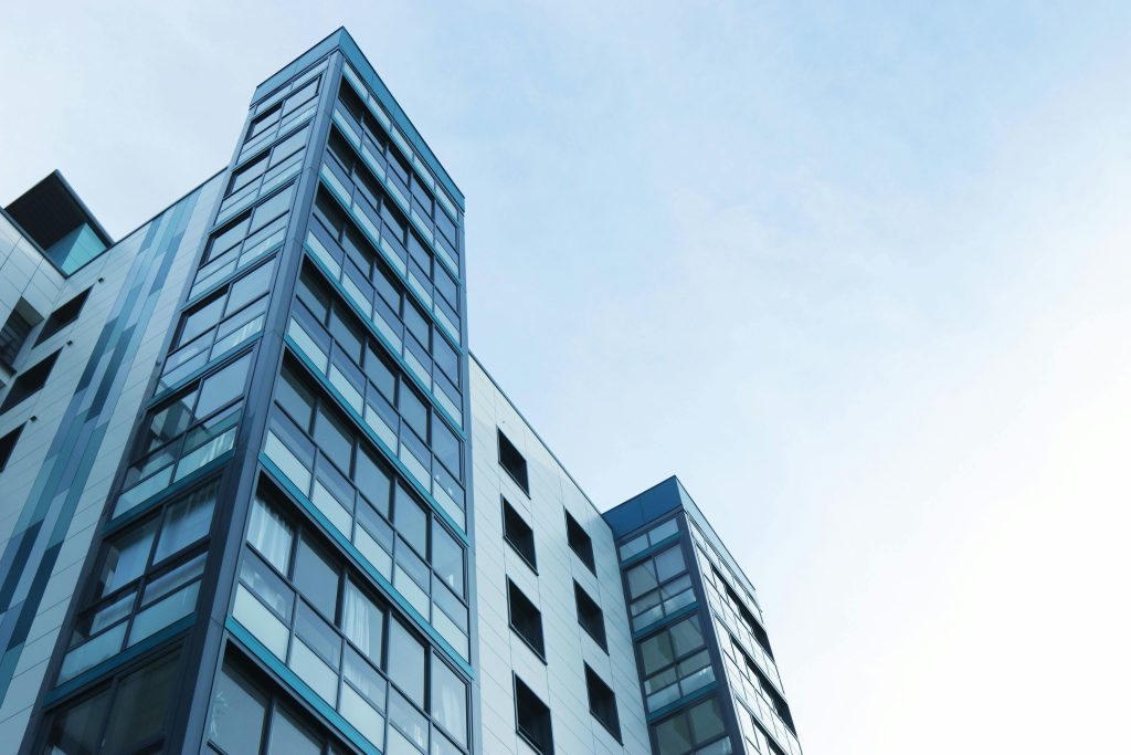 The modern high-rise building, home to various departments, boasts reflective glass windows against a clear blue sky. Viewed from a low angle, its facade showcases geometric patterns that highlight the contemporary architectural design.