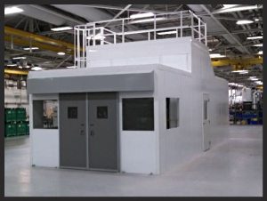 A large, white modular equipment enclosure with gray doors and windows sits in a spacious warehouse. The structure boasts a flat roof with railing, while the ceiling displays exposed metal beams. Green storage bins are visible in the background.
