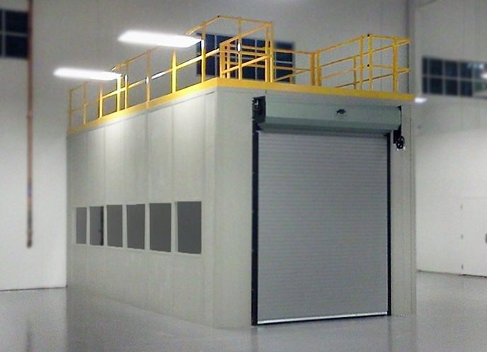A large industrial enclosure with a gray roll-up door built for secure equipment storage. The structure has multiple rectangular windows and a yellow safety railing on top. It is situated in a spacious, well-lit interior space with a smooth, glossy floor.