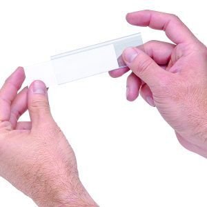 Two hands hold a clear microscope slide against a white background, fingers grasping gently on either side. It demonstrates precision akin to Quantum Label Holders with laser label inserts (25 per pack), underscoring meticulous attention to detail.