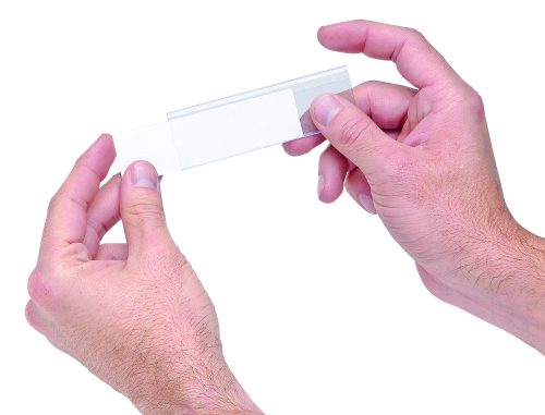 Two hands hold a clear microscope slide against a white background, fingers grasping gently on either side. It demonstrates precision akin to Quantum Label Holders with laser label inserts (25 per pack), underscoring meticulous attention to detail.