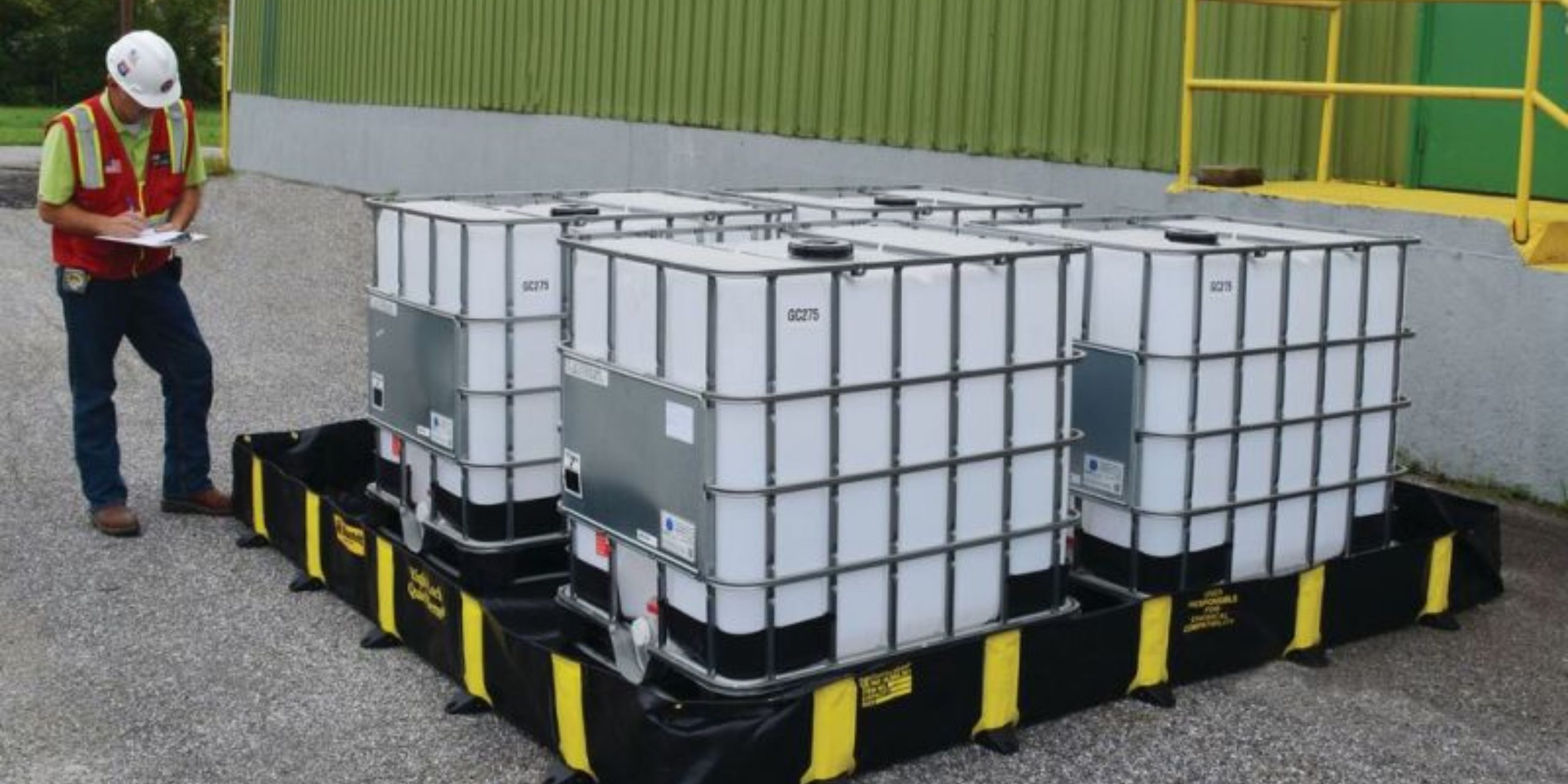 A worker in a safety vest and helmet inspects four large, white industrial containers on a spill containment pallet outside a green industrial building. The containers, secured with metal cages and labels, highlight advanced spill control measures.