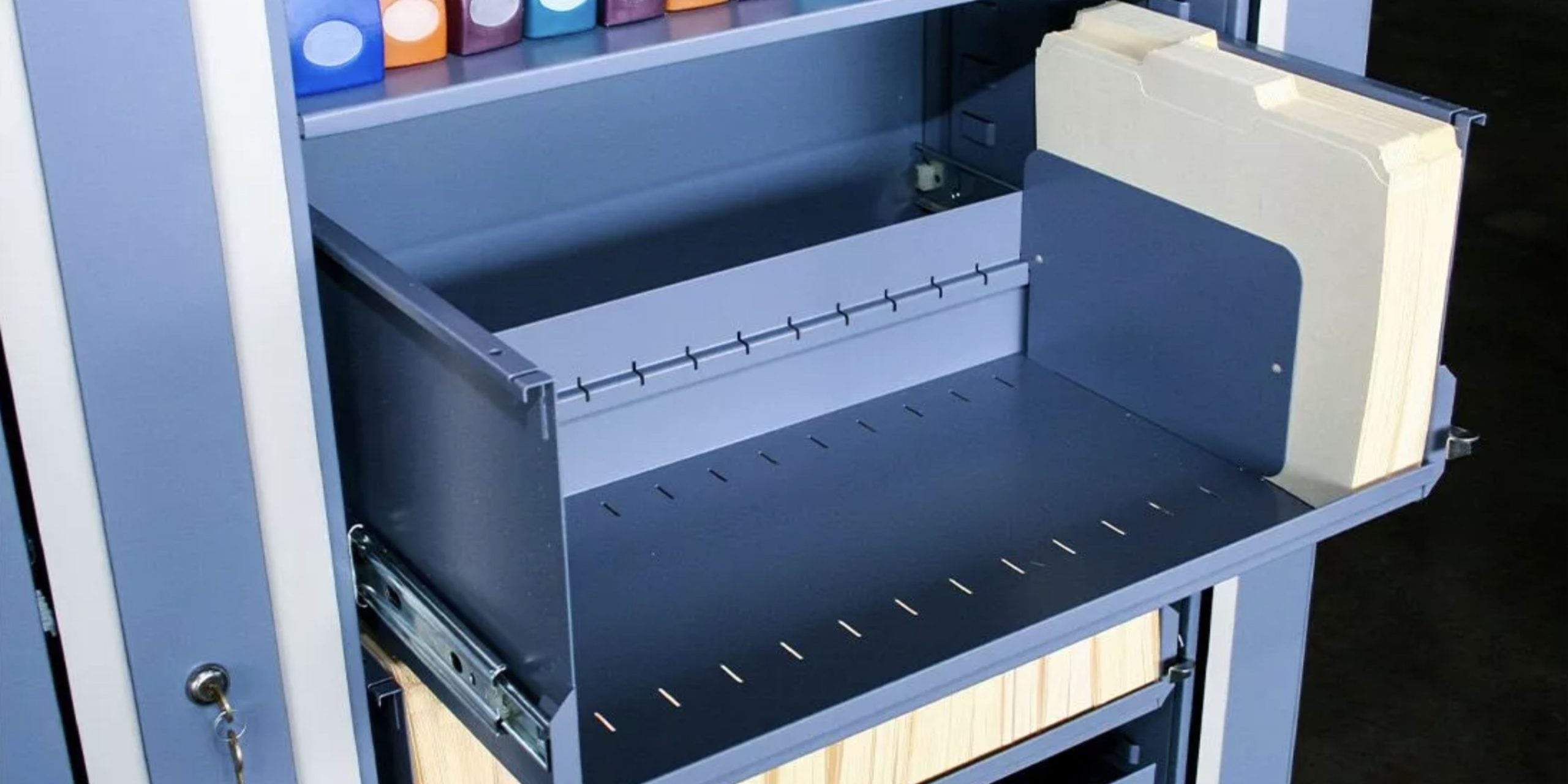 An open Times-2 Cabinet reveals a pull-out drawer lined with empty hanging folders. Above, vibrant binders perch on shelves. The unit is elegantly painted in shades of blue and gray, blending functionality with a splash of color.