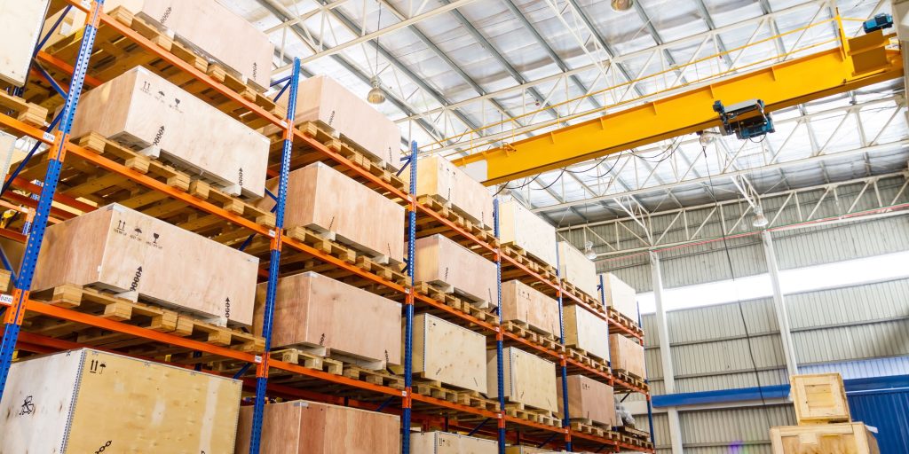 The warehouse interior showcases high stacks of wooden crates on robust orange and blue metal shelves, highlighting the importance of pallet rack safety features. A yellow overhead crane is suspended above, enhancing the spaciousness with its high ceiling and large, light-flooded windows.