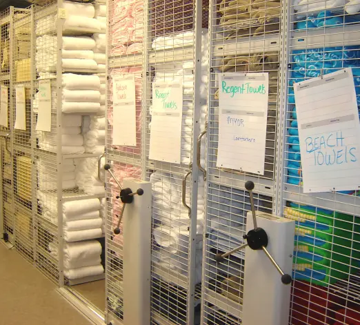 A storage room features E Z Rect Mobile Shelving Systems with metal shelves holding stacks of folded towels in various colors. Each section is labeled, including signs for Regent Towels and Beach Towels, and the shelves have a wheel for easy adjustment.