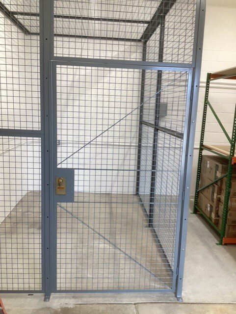 A sturdy security cage with a door, crafted from wire mesh and a steel frame, stands in a room with white brick walls. The door features a metal lock plate for added security. To the right, shelves hold cardboard boxes. The concrete floor completes this secure storage solution.