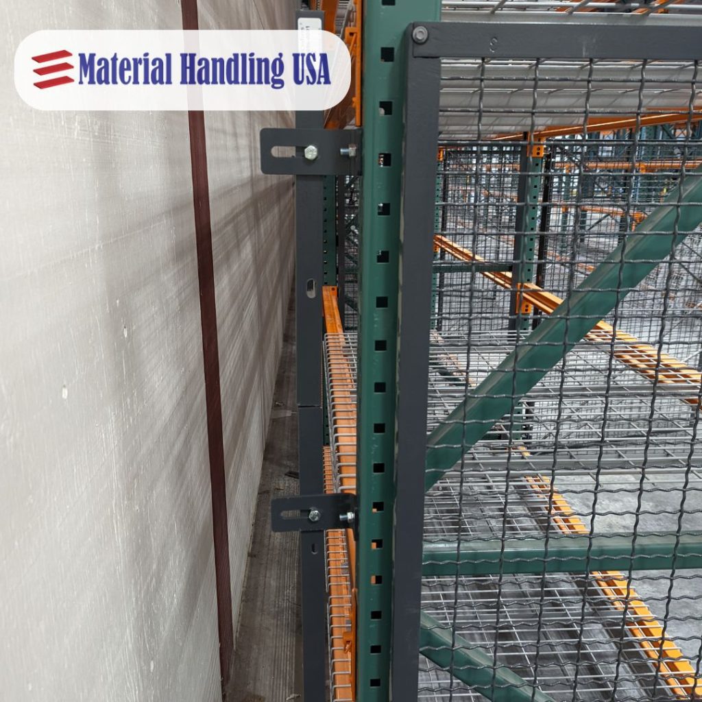 A heavy-duty warehouse shelving unit with metal wire mesh shelves and pallet rack backing is installed against a wall. A logo reading Material Handling USA is visible in the top left corner. Orange and green components support the structure, ensuring safety and durability.