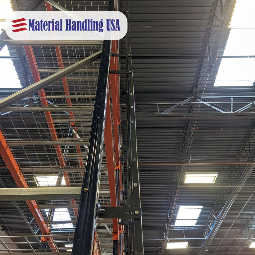 View of a warehouse interior with tall metal racks extending toward the ceiling, secured by rack safety barriers. The roof features industrial lighting and skylights. A logo in the top left corner reads Material Handling USA.