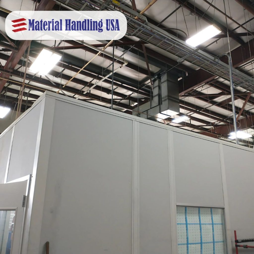 Interior of a warehouse with industrial-style lighting and steel beams. A large white structure with a partially open door is in view. Overhead, cables and ductwork are visible. A Material Handling USA sign is in the top left corner.