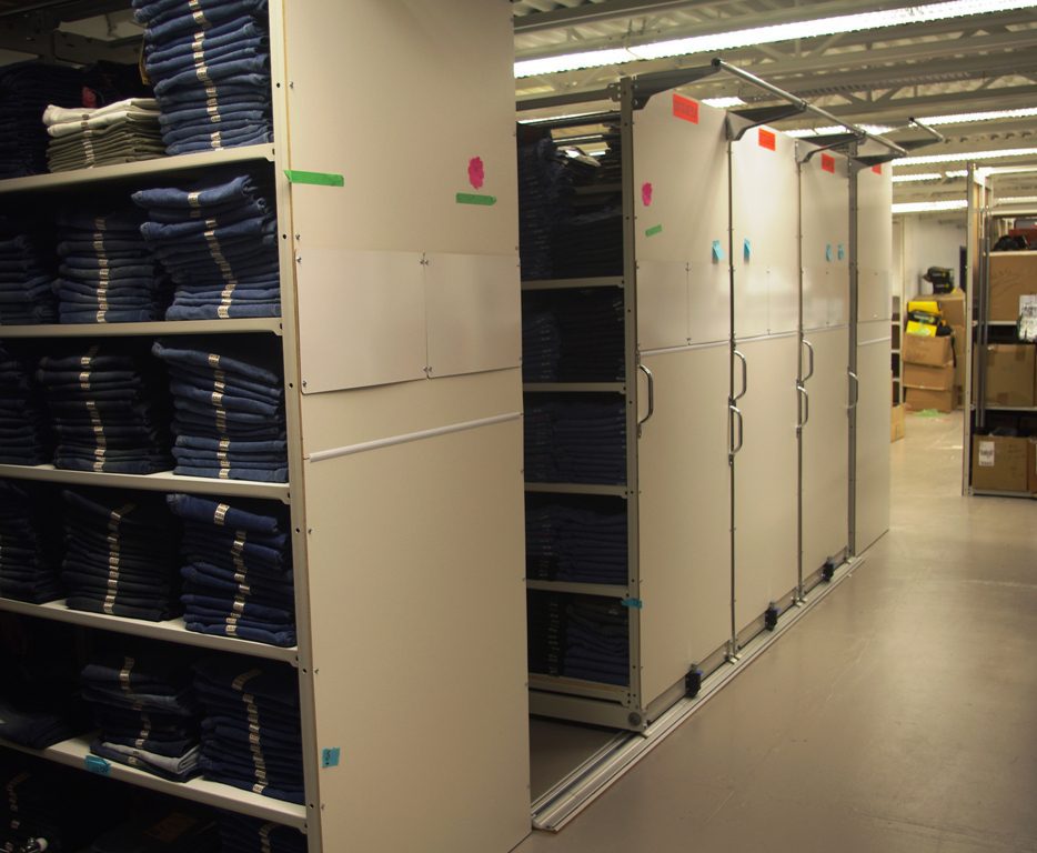 Shelving units filled with neatly stacked blue jeans in a storage room feature E Z Rect Mobile Shelving Systems. The shelves glide smoothly on tracks to form aisles. The room is well-lit, with colorful labels adding a vibrant touch.