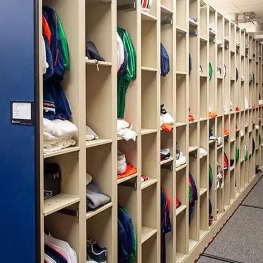 A row of vertical wooden cubbies filled with neatly organized clothing and shoes, showcasing various colors. The orderly setup suggests its part of a locker room with Durable Lockers for Athletics. A blue door is visible on the left side, complementing this sports facility space.