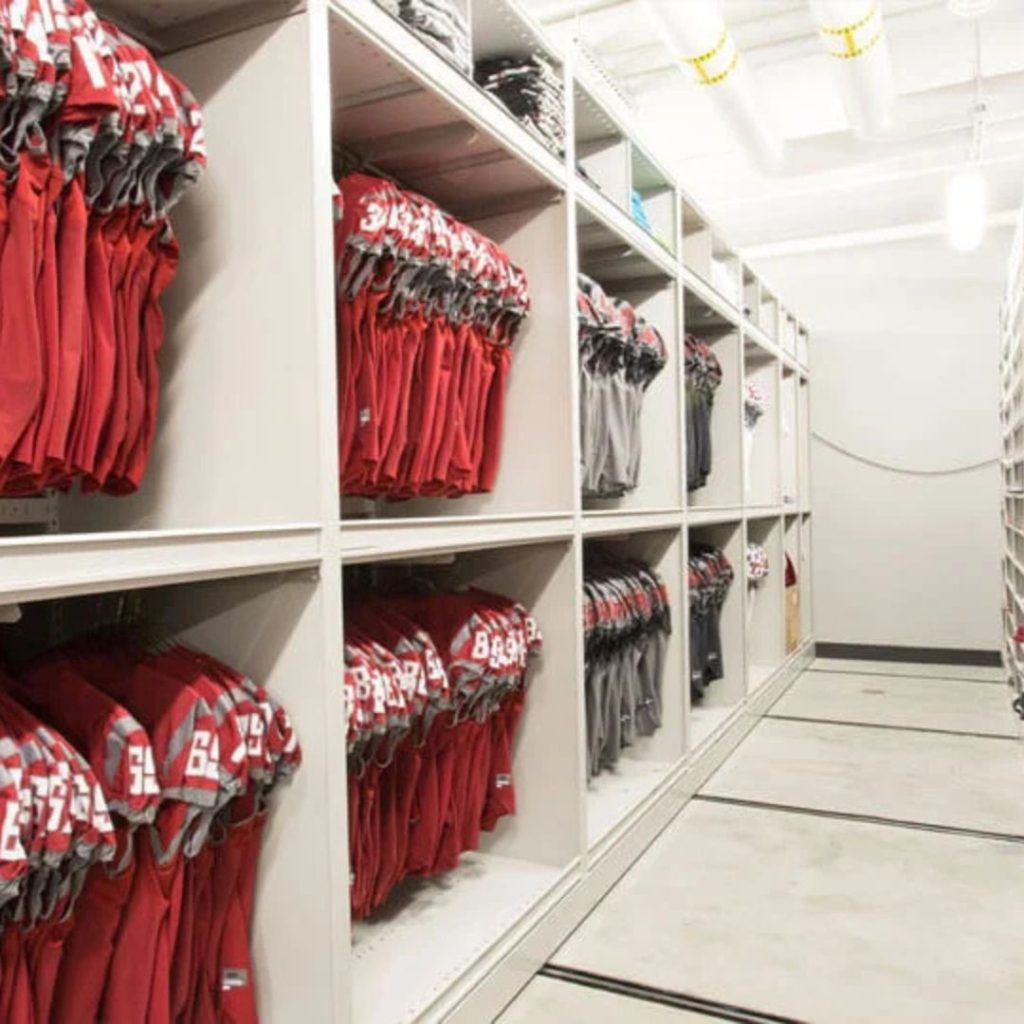 Shelves filled with red and gray sports jerseys, each emblazoned with the number 3 on the shoulders, line a narrow Athletic Storage room with white walls and overhead lighting. This efficient Sports Equipment Storage system showcases organization at its best.