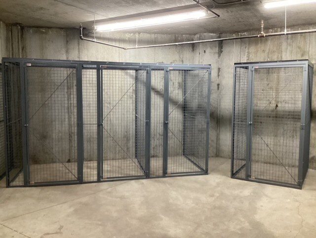 Metal storage cages in a concrete room, with shelves holding boxes and visible pipes in some units