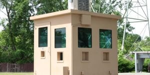 A tan, rectangular guard shack with large windows on all sides is situated outdoors. It is surrounded by trees, a fence, and metal structures, ensuring security. An air conditioning unit is mounted on the roof.
