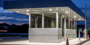 A modern, illuminated guard shack at night with reflective surfaces. The security booth is surrounded by traffic cones, and a barrier arm is visible on the right. The sky is dark blue with scattered clouds in the background.