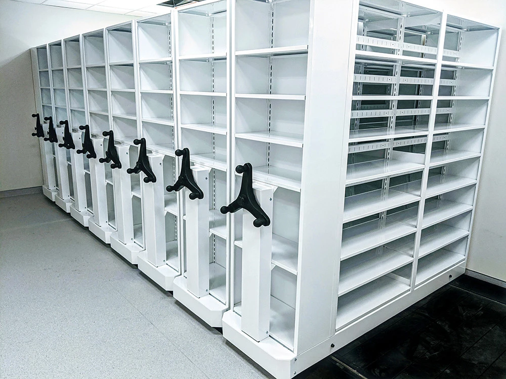 A row of white mobile shelving units with black rotating handles stands ready, showcasing Shelving Solutions for high-density storage. These empty shelves are perfectly aligned in a well-lit room with a light gray floor, embodying top-tier Space Optimization Solutions.
