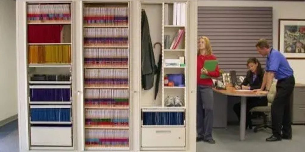 Office scene with two open cabinets full of color-coded files. Two people stand nearby, with one holding a green folder. Another person sits at a desk, focused on paperwork. A framed picture hangs on the wall.